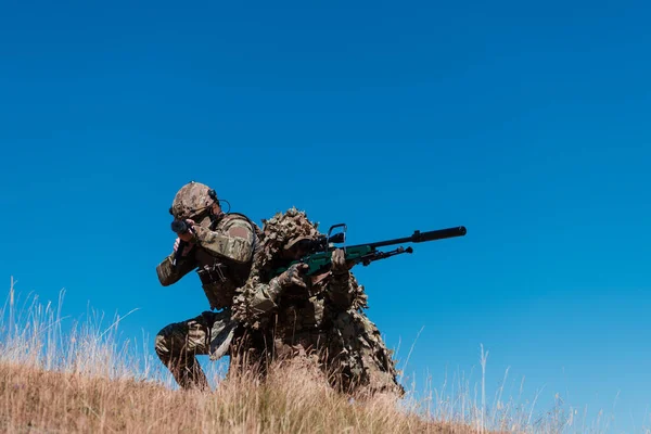 A sniper team squad of soldiers is going undercover. Sniper assistant and team leader walking and aiming in nature with yellow grass and blue sky. Tactical camouflage uniform. Hi quality stock photo.