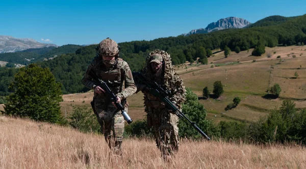 A sniper team squad of soldiers is going undercover. Sniper assistant and team leader walking and aiming in nature with yellow grass and blue sky. Tactical camouflage uniform. Hi quality stock photo.