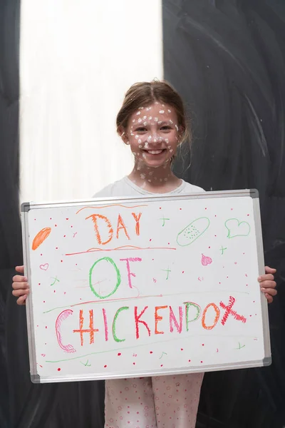 A little schoolgirl with chickenpox draws a message on the whiteboard in the kids room, and antiseptic cream is applied to the face and body. Chalkboard background. High quality photo