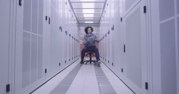 在现代云计算虚拟数据中心超级计算机的服务器室内从事平板计算机工作的It工程师或数据科学专家 嬉皮士身穿休闲装 坐在椅子上 — 图库视频影像
