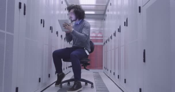 Itエンジニアやデータサイエンスの専門家は 現代のクラウドコンピューティング仮想データセンタースーパーコンピュータのサーバールームでタブレットコンピュータに取り組んでいます カジュアルな服を着て椅子に座っているヒップスター — ストック動画