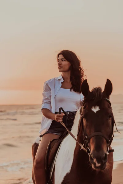 A woman in summer clothes enjoys riding a horse on a beautiful sandy beach at sunset. Selective focus. High quality photo