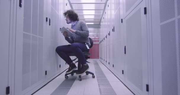 Itエンジニアやデータサイエンスの専門家は 現代のクラウドコンピューティング仮想データセンタースーパーコンピュータのサーバールームでタブレットコンピュータに取り組んでいます カジュアルな服を着て椅子に座っているヒップスター — ストック動画