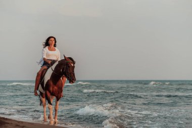 Yaz kıyafetleri içinde bir kadın gün batımında kumlu güzel bir sahilde ata binmekten hoşlanır. Seçici odaklanma. Yüksek kalite fotoğraf