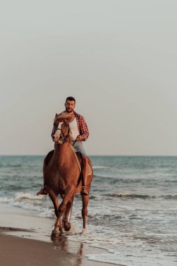 Yaz kıyafetleri içinde modern bir adam gün batımında kumlu güzel bir sahilde ata binmekten hoşlanır. Seçici odaklanma. Yüksek kalite fotoğraf