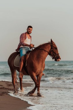 Yaz kıyafetleri içinde modern bir adam gün batımında kumlu güzel bir sahilde ata binmekten hoşlanır. Seçici odaklanma. Yüksek kalite fotoğraf