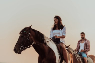 Gün batımında kumlu bir sahilde ata binen yaz kıyafetleri içinde sevgi dolu bir çift. Arka planda deniz ve gün batımı. Seçici odaklanma. Yüksek kalite fotoğraf
