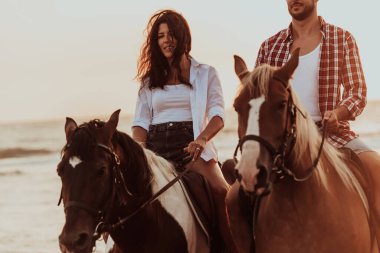 Gün batımında kumlu bir sahilde ata binen yaz kıyafetleri içinde sevgi dolu bir çift. Arka planda deniz ve gün batımı. Seçici odaklanma. Yüksek kalite fotoğraf