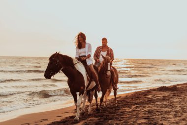 Gün batımında kumlu bir sahilde ata binen yaz kıyafetleri içinde sevgi dolu bir çift. Arka planda deniz ve gün batımı. Seçici odaklanma. Yüksek kalite fotoğraf