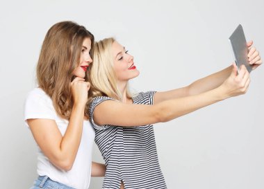 Dijital tabletle selfie çeken iki bayan arkadaş. Stüdyo, gri arka planda çekim yapıyor.