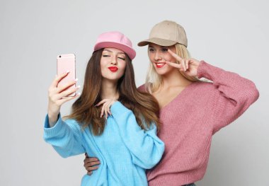 Kazak ve şapka giymiş iki kadının portresi. Gri arka planda durup selfie çekiyorlar.
