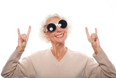 Büyük güneş gözlüğü takan yaşlı bir kadın izole edilmiş beyaz arka planda durup çılgın bir ifadeyle el kaldırarak rock sembolü yapıyor. Müzik yıldızı. Ağır konsept.