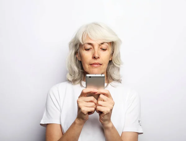 Portrait of old smiling lady looking at screen of mobile phone over grey background