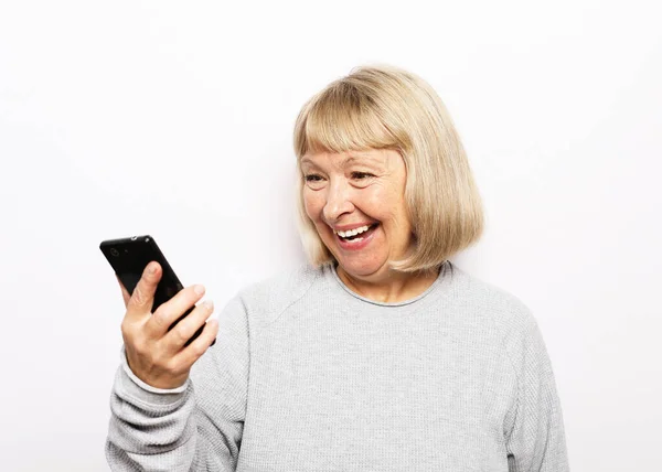 穿着休闲装的可爱的老太太 站在白色的背景上 用现代智能手机开心地微笑着 看着相机 生活方式和技术概念 — 图库照片