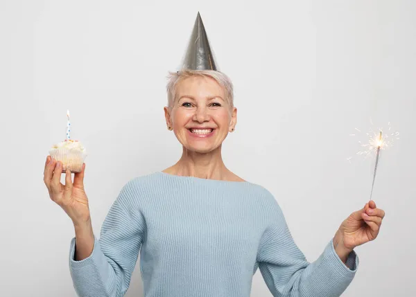 Happy middle aged female wearing conical hat celebrating birthday with cupcake with candle and sparkler — Stock Photo, Image