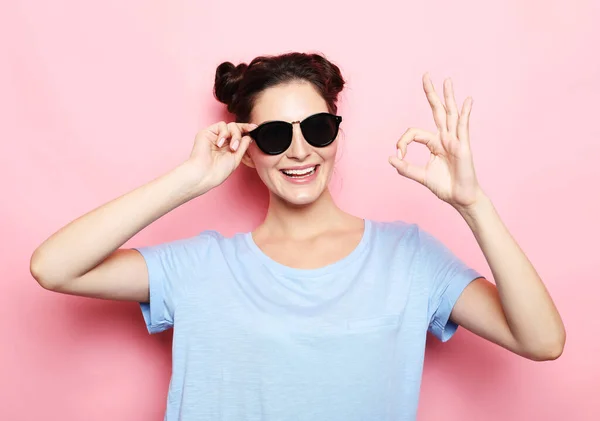身穿蓝色T恤 戴着太阳镜的年轻快乐的黑发女人在粉红的背景上显示出可以接受的标志 — 图库照片