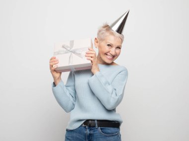 Neşeli, pozitif, mutlu, mavi kazak giyen, parti şapkası takan, beyaz gri arka planda izole tatil havasının tadını çıkaran yaşlı kadının portresi..