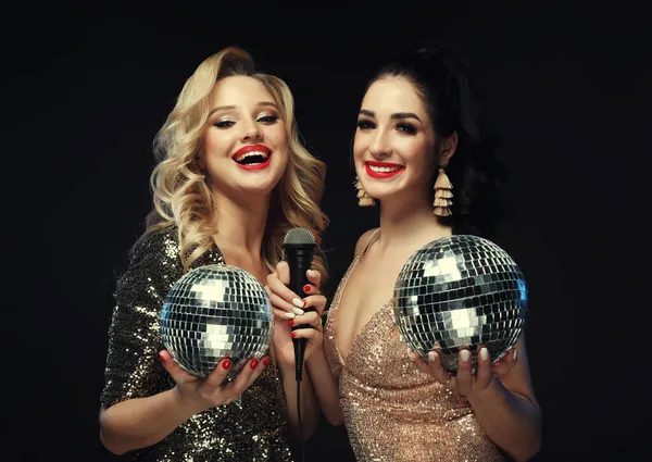 Dwie piękne kobiety, blondynka i brunetka w lśniących sukienkach, śpiewają z mikrofonem, trzymając kulę dyskotekową — Zdjęcie stockowe