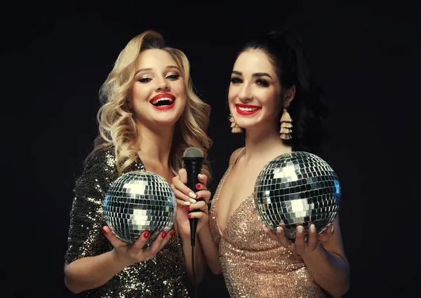 İki güzel kadın, bir sarışın ve parlak elbiseli bir esmer, mikrofonla şarkı söylüyorlar, disko topu tutuyorlar.