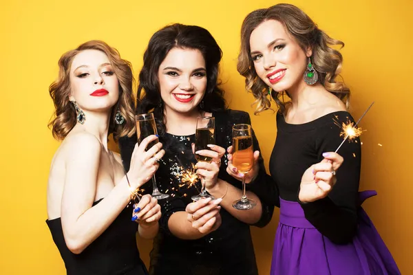 Tre unga kvinnor med champagne glasögon och tomtebloss över gul bakgrund — Stockfoto