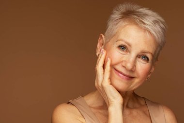 Elleri yüzlerinde gülümseyen olgun bir kadının güzel portresi. Yaşlanma karşıtı tedaviden sonra kendini zinde hisseden mutlu son sınıf kadınının yakın çekimi. Gülümseyen güzel kusursuz ciltli kameraya bakıyor..