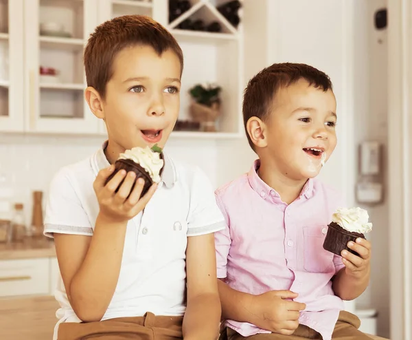 Funny two little brothers are sitting together at the kitchen table and eating cupcakes. — Stock Photo, Image