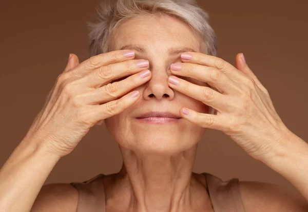 Portrait of beautiful senior woman touching her perfect skin andclosed eyes. Closeup face of mature woman with wrinkles massaging face isolated over brown background. Aging process concept.