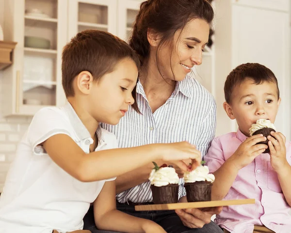 Familia feliz en la cocina. Mamá y dos niños pequeños, hermanos, están comiendo cupcakes caseros. —  Fotos de Stock