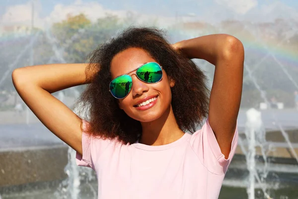 Jong Afrikaans vrouwtje met zonnebril en roze t-shirt poserend bij de fontein — Stockfoto