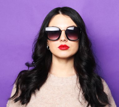 Uzun kıvırcık saçlı çekici bir kadın moda güneş gözlüğü takıyor ve kameraya bakıyor.