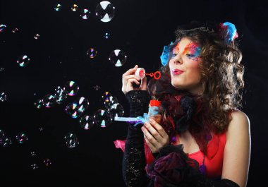 Yaratıcı makyajlı, sabun köpüğü üfleyen modaya uygun genç bir kadın. Bebek stili. Parlak elbise. Siyah arkaplan.