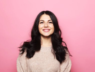 Genç, güzel, kıvırcık saçlı, pembe süveter giyen, yüzünde mutlu ve havalı bir gülümseme olan esmer kadın..