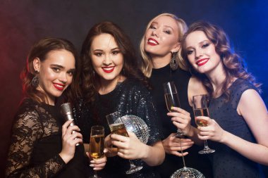 Bir grup parti kadını flütleri köpüklü şarapla tokuşturuyor ve ellerinde disko topları ve mikrofon tutuyorlar..