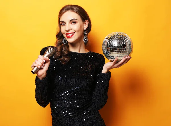 Young woman in evening dress holding microphone and disco ball. Brighrt make up and Wavy hairstyle. — Stock Photo, Image