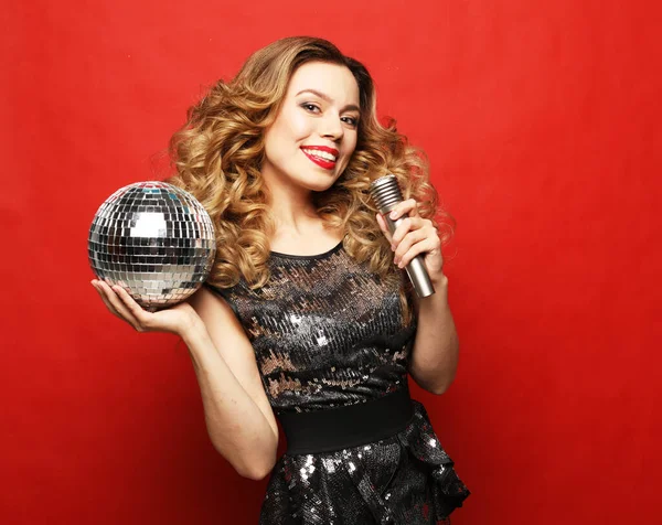 Junge blonde Frau mit langen welligen Haaren im Abendkleid mit Mikrofon und Discokugel — Stockfoto