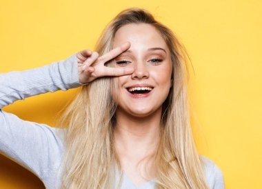 Gülümseyen mutlu sarışın bir kadının portresi zafer işareti gösteriyor ve sarı arkaplanda izole edilmiş kameraya bakıyor.