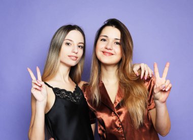 İki güzel kadın pijama giyip sarılıyorlar ve mor arka planda barış işareti gösteriyorlar..
