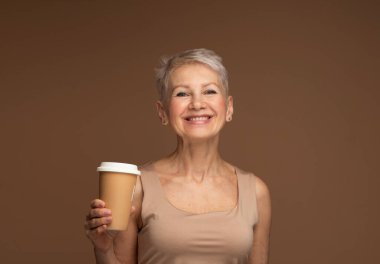 Yaşlı, gülümseyen, modaya uygun, mutlu, beyaz bir kadın kahverengi arka planda izole olmak için paket bej kahve taşıyor..