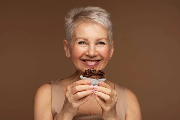 髪の短い年配の女性は手にチョコレートカップケーキを持っている 茶色の背景に肖像画 — ストック写真