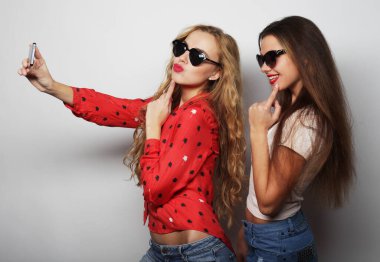 Arkadaşlık, insanlar ve teknoloji kavramı. Akıllı telefonlu iki mutlu genç kadın beyaz arka planda selfie çekiyor.