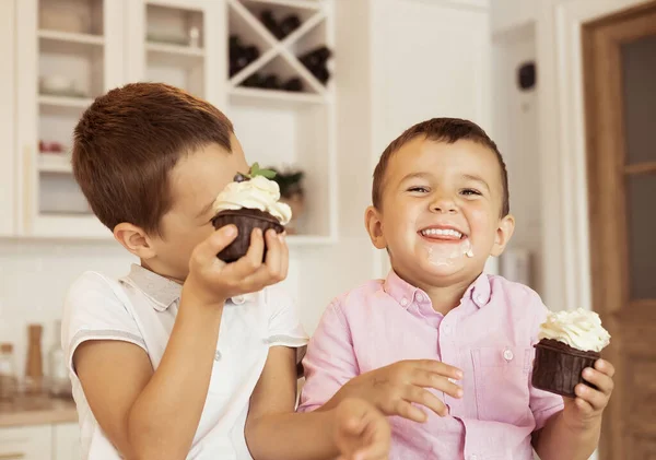 Funny two brother male kids are sitting together at the kitchen table and eating cupcakes. — Stock Photo, Image