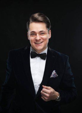 Smokinli şık bir genç elinde mikrofonla karanlık bir arka planda dumanla poz veriyor.