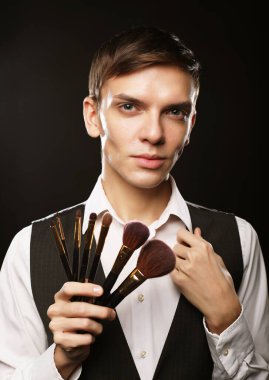 Genç erkek makyaj sanatçısı koyu bir arka planda makyaj fırçasıyla poz veriyor.