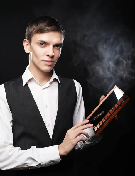 Male make-up artist staying on dark background, holding professional brush and shadows palette — Stock Photo, Image