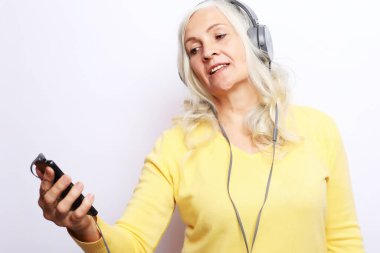Sarı kazak giyen neşeli yaşlı kadın beyaz arka planda kulaklıkla müzik dinliyor..