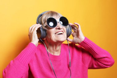 Pembe kazak ve büyük güneş gözlüğü takan neşeli yaşlı kadın sarı arka planda kulaklıklı müzik dinliyor..