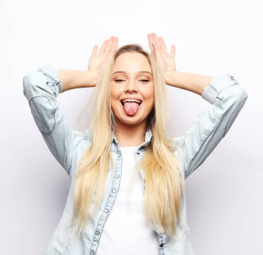 Yaşam tarzı, duygu ve vücut dili kavramı: Beyaz arka plan üzerinde gündelik giysiler giyen genç bir kadın boynuzları üzerinde parmaklarıyla komik hareketler yapıyor.