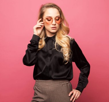 Güzel, genç, sarışın bir kadın pembe arka planda şık kıyafetler içinde poz veriyor..