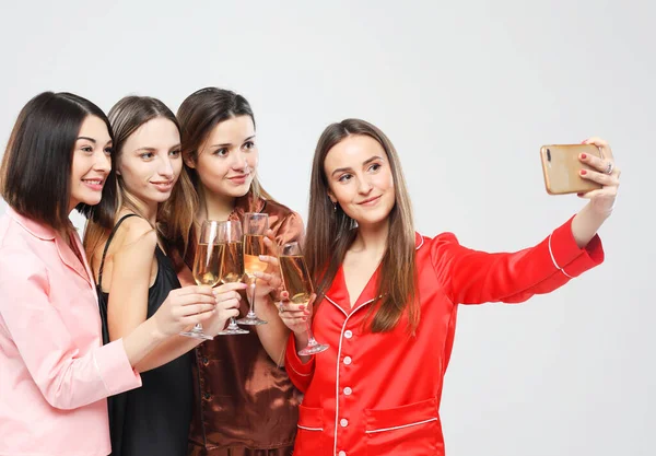 happy friends women wearing pajamas with smartphone taking selfie, drinking wine and having fun, party time.
