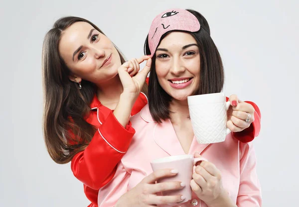 Due belle giovani donne vestite in pigiama che tengono tazze, abbracciano e sorridono — Foto Stock